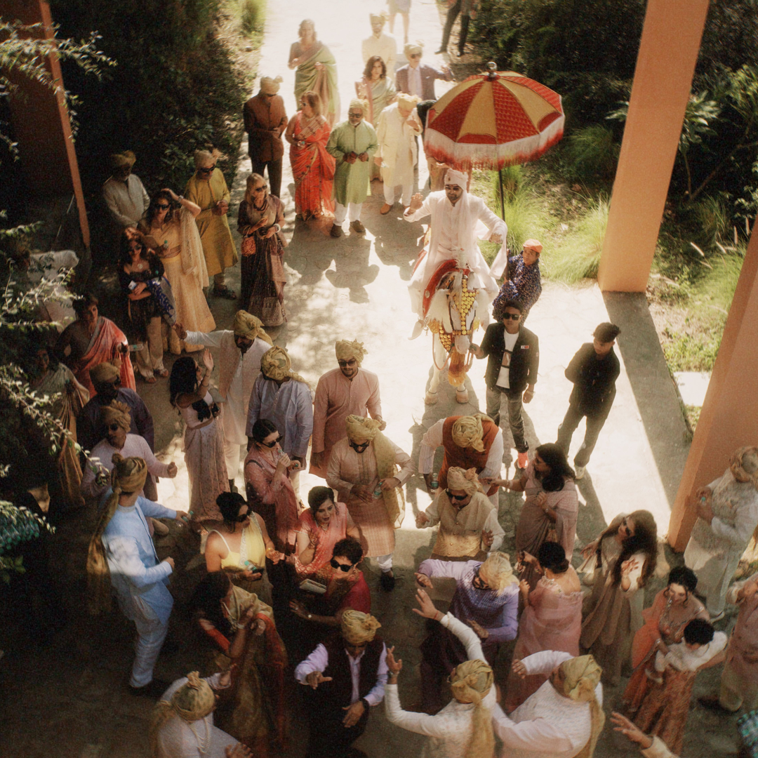 Wedding guests in colourful outfits gathered under parasols at an outdoor procession